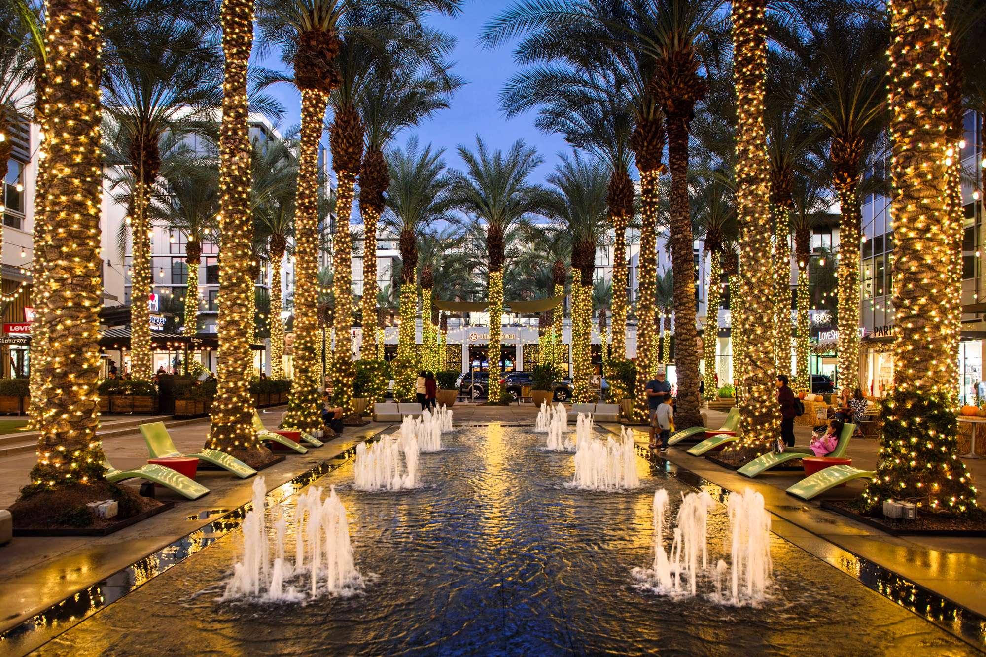 The Scottsdale Quarter open-air shopping center, showing the plaza fountain & palm trees wrapped in seasonal holiday lights.