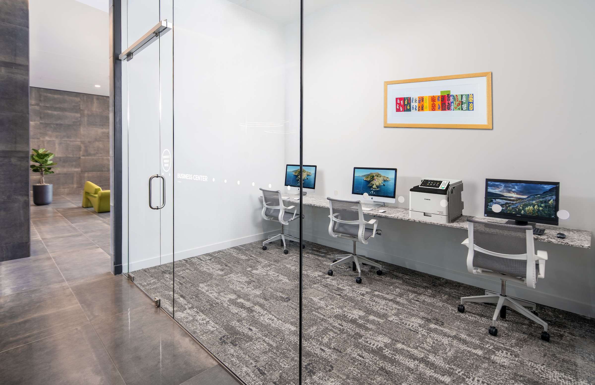 Business station with Apple desktop computers & ergonomic mesh office chairs within a glass-walled room at Optima Kierland.