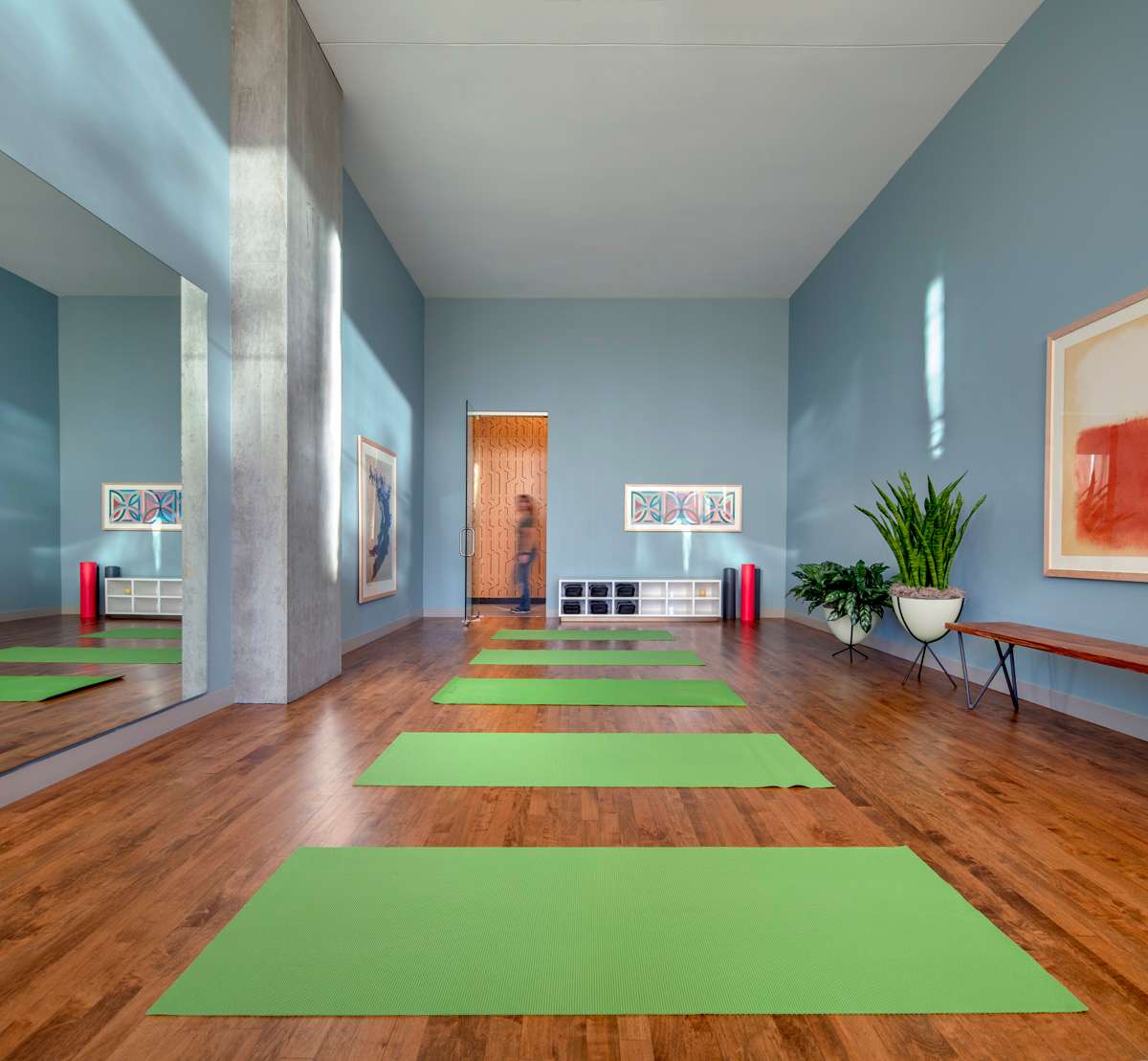 A yoga studio with rich hardwood flooring, natural lighting, evenly spaced bright green yoga mats, & pale blue walls.