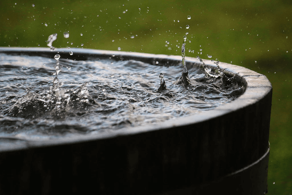 Raindrops landing on the surface of a rainwater harvesting system, storing the water in a barrel for later use.