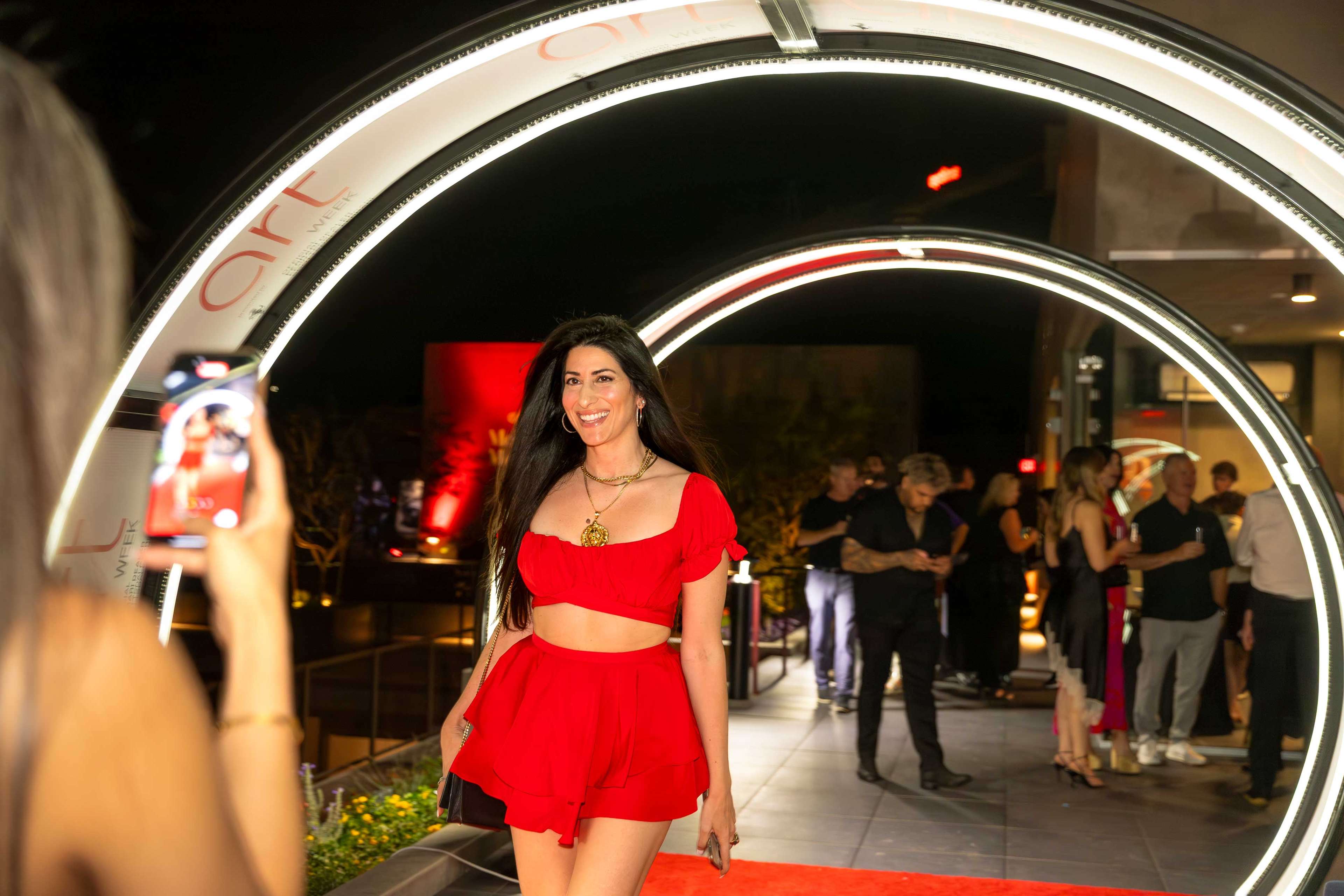 A woman attending an evening event, wearing a red two-piece set with a draped skirt, and holding a black handbag.